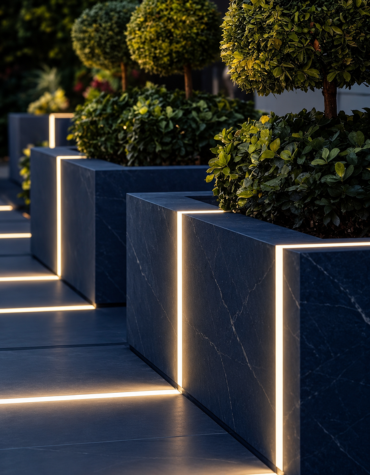 Modern navy blue marble-finish planter boxes with integrated warm white LED profiles illuminating the edges, arranged along a contemporary outdoor walkway with green shrubs and ornamental trees.