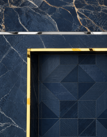 Close-up of navy blue marble wall cladding with natural white and gold veining, accented by a sleek stainless steel horizontal profile and polished gold edging around a geometric dark blue tiled panel.