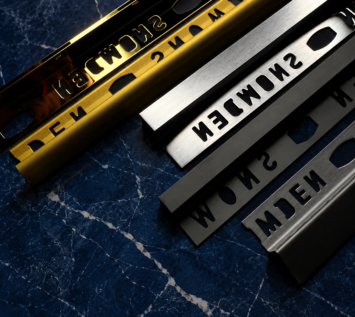 Close-up of gold, silver, and black metal tile trim profiles arranged diagonally on a dark blue marble surface, showcasing premium finishes for modern architectural and interior design applications.